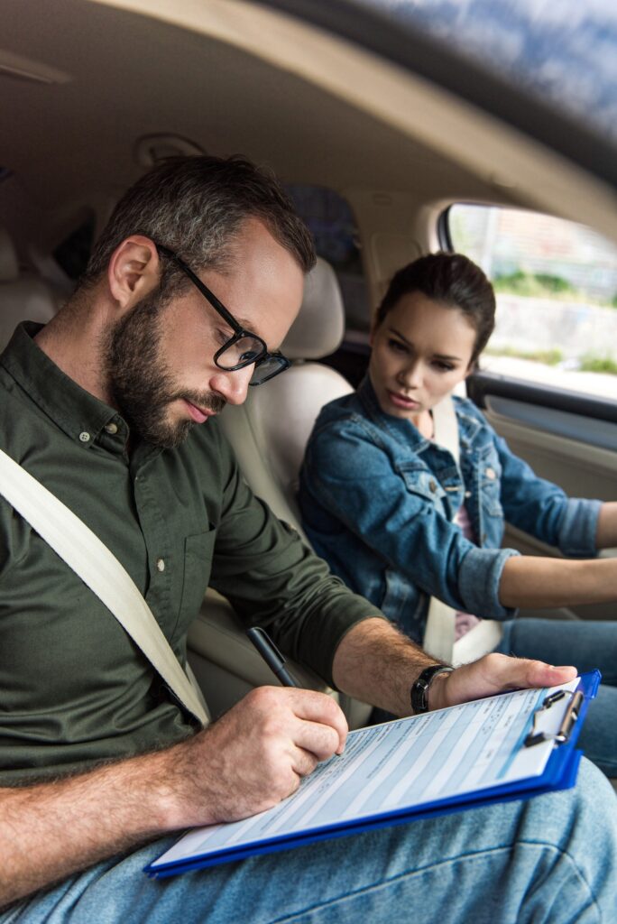 Fahrlehrer sitzt im Auto und notiert etwas in seinen Unterlagen. Die Fahrschülerin sitzt neben ihm und schaut, was er schreibt. Sie absolviert einen 2 Phasen Kurs.